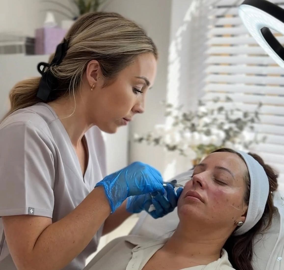 Amy in grey scrubs treating a client's cheek beside the window